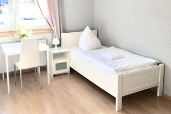 Helles und schlicht eingerichtetes Einzelzimmer im G&auml;stehaus Odin mit wei&szlig;em Bett, Tisch, Stuhl und Fenster mit Vorhang.