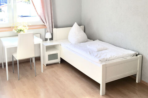Helles und schlicht eingerichtetes Einzelzimmer im G&auml;stehaus Odin mit wei&szlig;em Bett, Tisch, Stuhl und Fenster mit Vorhang.