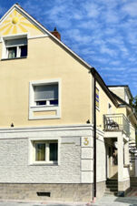 Fassade des G&auml;stehaus Odin mit heller Wand, wei&szlig;en Fenstern und einer goldgelben Sonnenzeichnung in der Dachspitze bei klarem Himmel.