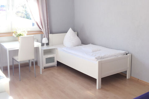 Helles Zimmer im G&auml;stehaus Odin mit einem Einzelbett, wei&szlig;em Schreibtisch, Stuhl und Fenster mit Gardinen.