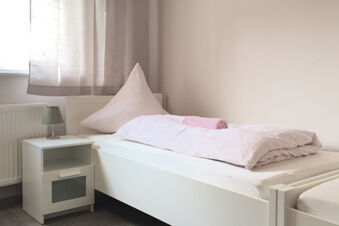 Hell eingerichtetes Einzelzimmer im G&auml;stehaus Odin mit einem wei&szlig;en Bett, rosa Bettw&auml;sche und einem Nachttisch mit Lampe.