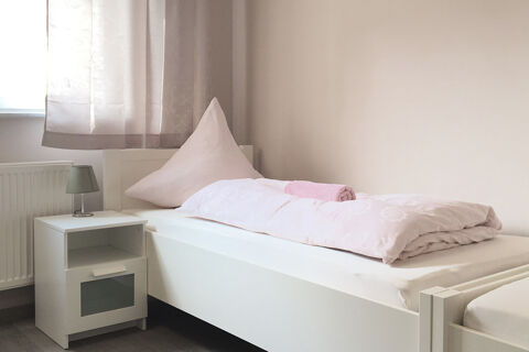 Hell eingerichtetes Einzelzimmer im G&auml;stehaus Odin mit einem wei&szlig;en Bett, rosa Bettw&auml;sche und einem Nachttisch mit Lampe.