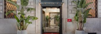 Entrance of Hotel NH Collection Roma Fori Imperiali with glass doors flanked by large potted plants and a red welcome mat inside