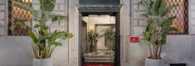 Entrance of Hotel NH Collection Roma Fori Imperiali with glass doors flanked by large potted plants and a red welcome mat inside