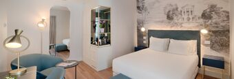 Bright hotel room at Hotel NH Collection Roma Fori Imperiali with white bed, blue armchair, wooden floor, and black and white wall art