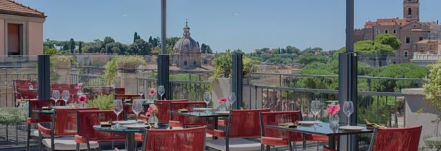 Outdoor dining area with red chairs and glass-railed terrace with city views at Hotel NH Collection Roma Fori Imperiali