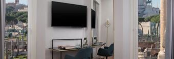 A hotel room at Hotel NH Collection Roma Fori Imperiali with a wall-mounted TV, desk with chair, glass water bottles, and a window view of Roman ruins