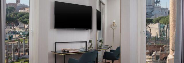 A hotel room at Hotel NH Collection Roma Fori Imperiali with a wall-mounted TV, desk with chair, glass water bottles, and a window view of Roman ruins