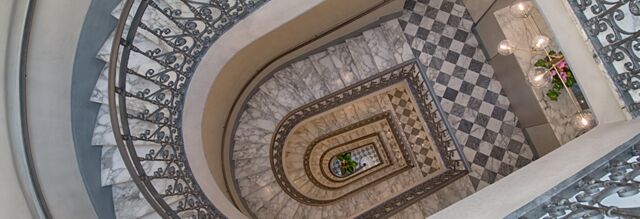 Spiral marble staircase with ornate iron railings and checkered tile floor at Hotel NH Collection Roma Fori Imperiali