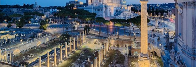 Ancient Roman ruins with columns and stone structures lit up at dusk near the Hotel NH Collection Roma Fori Imperiali
