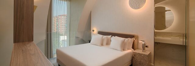 A modern hotel room at NH Collection Milano CityLife with a large bed, white pillows, stone nightstand, and a glass partition bathroom