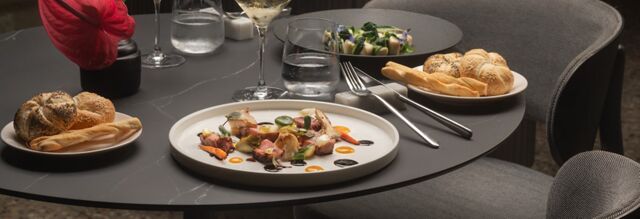 A gray round table at NH Collection Milano CityLife with a plated gourmet dish, two bread plates, a glass of wine, and water glass