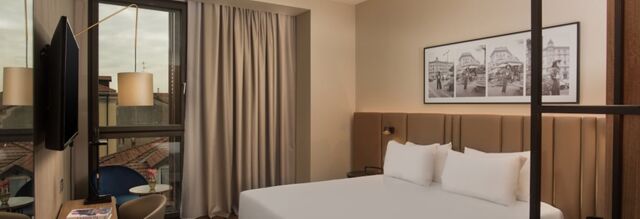 Hotel NH Milano Corso Buenos Aires room with a large bed, three pillows, beige padded headboard, and window with curtains and a balcony chair