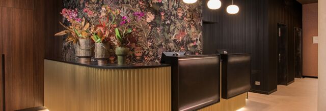 Reception area with a ribbed golden counter, floral wallpaper, flower arrangements, and dark wood paneling at Hotel NH Milano Corso Buenos Aires