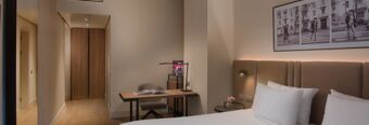 Hotel NH Milano Corso Buenos Aires room with a white bed, brown headboard, bedside table, desk with chair, and framed black and white pictures