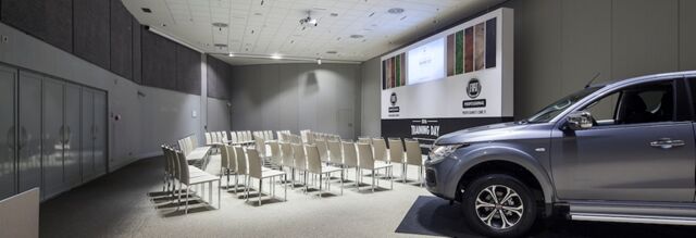 A conference room at NH Milano Congress Centre with rows of white chairs facing a presentation screen and a gray pickup truck parked nearby