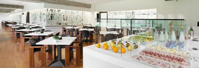 Bright modern dining area with wooden floors, white tables, black chairs, and a glass-covered buffet at NH Milano Congress Centre