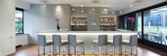 Modern bar area with stools, illuminated counter, shelves of drinks, purple orchids, and large windows at NH Milano Congress Centre