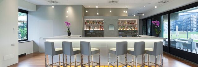 Modern bar area with stools, illuminated counter, shelves of drinks, purple orchids, and large windows at NH Milano Congress Centre