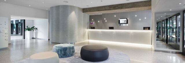 Reception area of NH Milano Congress Centre with a white illuminated desk, three round ottomans, and glass entrance doors visible