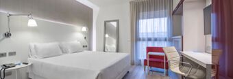 A modern hotel room at NH Milano Congress Centre with a large bed, white linens, wall lamps, a red chair, desk, and window with curtains