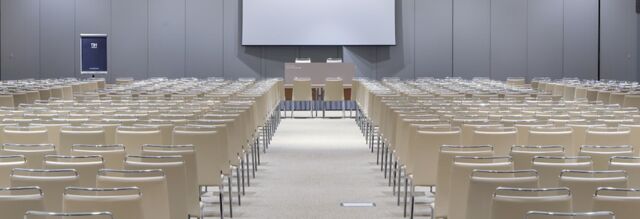 Empty conference room at NH Milano Congress Centre with rows of beige chairs facing a stage and projector screen