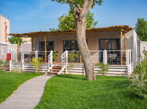 A wooden patio with chairs and tables behind a white fence with steps, green grass and a tree in front at Rimini Family Resort