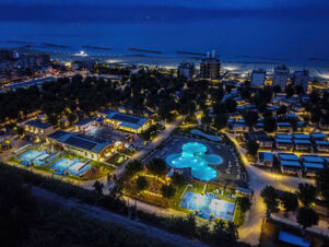 Aerial night view of Rimini Family Resort with illuminated swimming pool, tennis courts, buildings, trees, beach, and calm sea in background