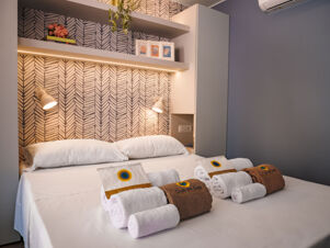 White bed with pillows and rolled towels placed on top, bedside lamps and shelves with decor in Rimini Family Resort room