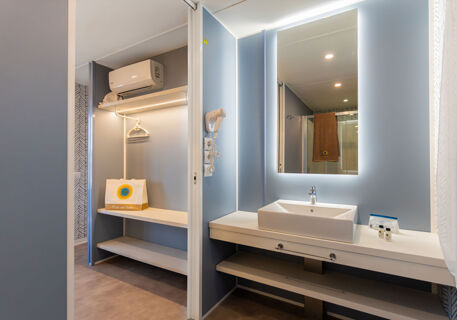 Modern bathroom and closet area with illuminated mirror, white sink, hairdryer, and branded items at Romagna Family Resort