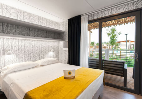 Double bed with white linens and yellow runner in a Romagna Family Resort room with patterned wall and patio door view outside