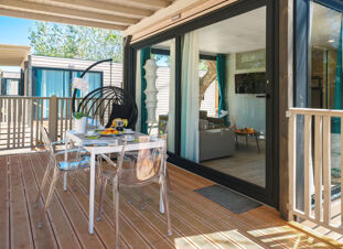 Outdoor wooden deck at Romagna Family Resort with a white table set for breakfast, transparent chairs, and a hanging wicker chair with a cushion