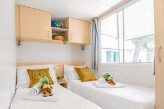 Two single beds with white bedding and yellow pillows with circular patterns inside a room at Riccione Easy Camping Village with a window and wooden cabinets above the beds
