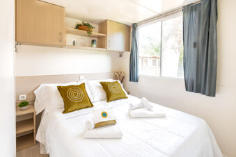Double bed with two green cushions and white linens under wooden cabinets next to a large window with blue curtains at Riccione Easy Camping Village