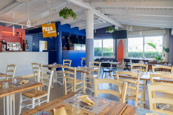 Interior of Riccione Easy Camping Village pizza restaurant with wooden tables, chairs, hanging plants, and a coffee machine area visible