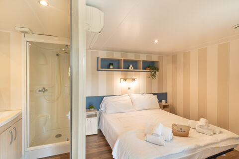 Bed with white linens and towels, small bedside tables, wall-mounted wooden shelves, and shower cabin in a Marina Family Resort room