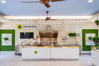 Chef serving food behind a glass counter in a kitchen area with Club del Sole logo at Marina Family Resort