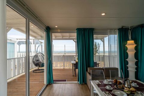 Indoor view of dining table with breakfast in front of large windows and doors opening to a wooden deck with teal curtains at Pini Boutique Resort