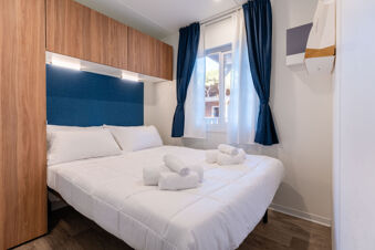 White bed with rolled towels, blue headboard, wooden overhead cabinets, window with blue and white curtains in Rivaverde Easy Camping Village