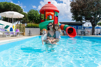 A woman with two young children in a swimming pool in front of a colorful playground slide at Adriatico Cervia Easy Camping Village
