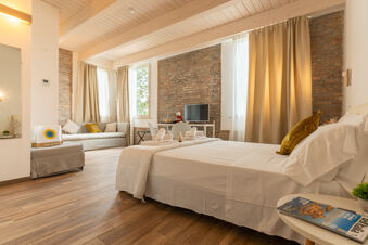 Hotel room at Vigna sul Mar Family Collection with a white bed, mustard pillows, a sofa, TV, wooden floors, and beige curtains
