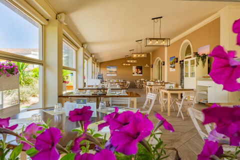 Indoor dining area at Vigna sul Mar Family Collection with wooden tables, white chairs, hanging lights, and large windows with flowers outside