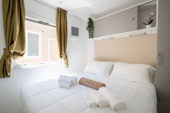 White bedding with towels folded on top, a window with olive curtains, overhead cabinets and plants in the La Risacca Family Collection room