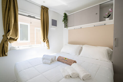 White bedding with towels folded on top, a window with olive curtains, overhead cabinets and plants in the La Risacca Family Collection room