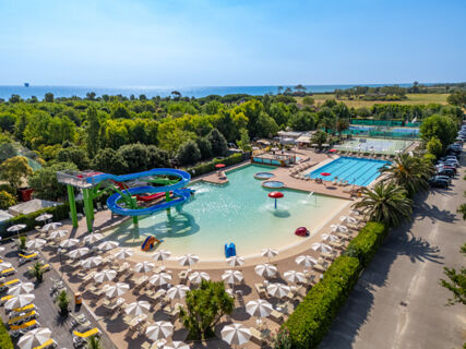 Outdoor water park with a large pool, water slides, sun umbrellas, tennis courts, and greenery near the sea at La Risacca Family Collection