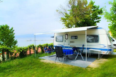 A white and blue caravan with an awning extended, outdoor table with a blue cover, and chairs on grass by a lake at Le Palme Lazise Family Collectio