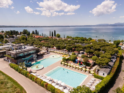 Aerial view of two swimming pools, water slides, lounge chairs, umbrellas, playground, and lake shore at Le Palme Lazise Family Collectio