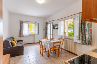 Dining area with a table set for breakfast, a gray sofa with yellow pillows, floral curtains, and open windows at Le Palme Lazise Family Collectio
