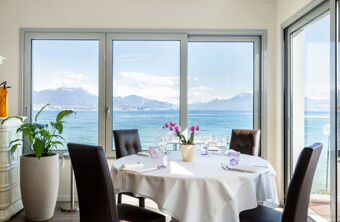 Dining table with white tablecloth, chairs, orchid flower, glassware, plants inside room overlooking sea and mountains at Desenzano Boutique Resor