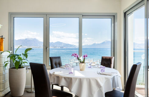 Dining table with white tablecloth, chairs, orchid flower, glassware, plants inside room overlooking sea and mountains at Desenzano Boutique Resor